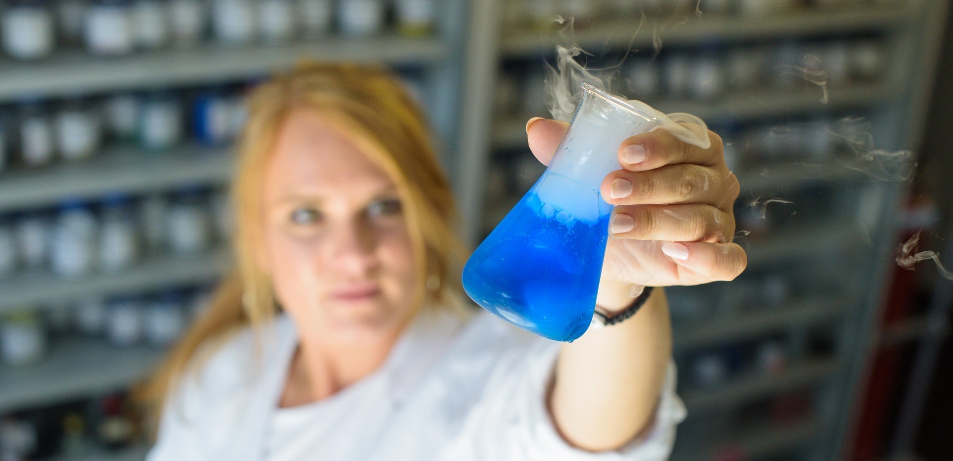 Studentin in einem Kittel hält ein Reagenzglas mit blauer Flüssigkeit hoch