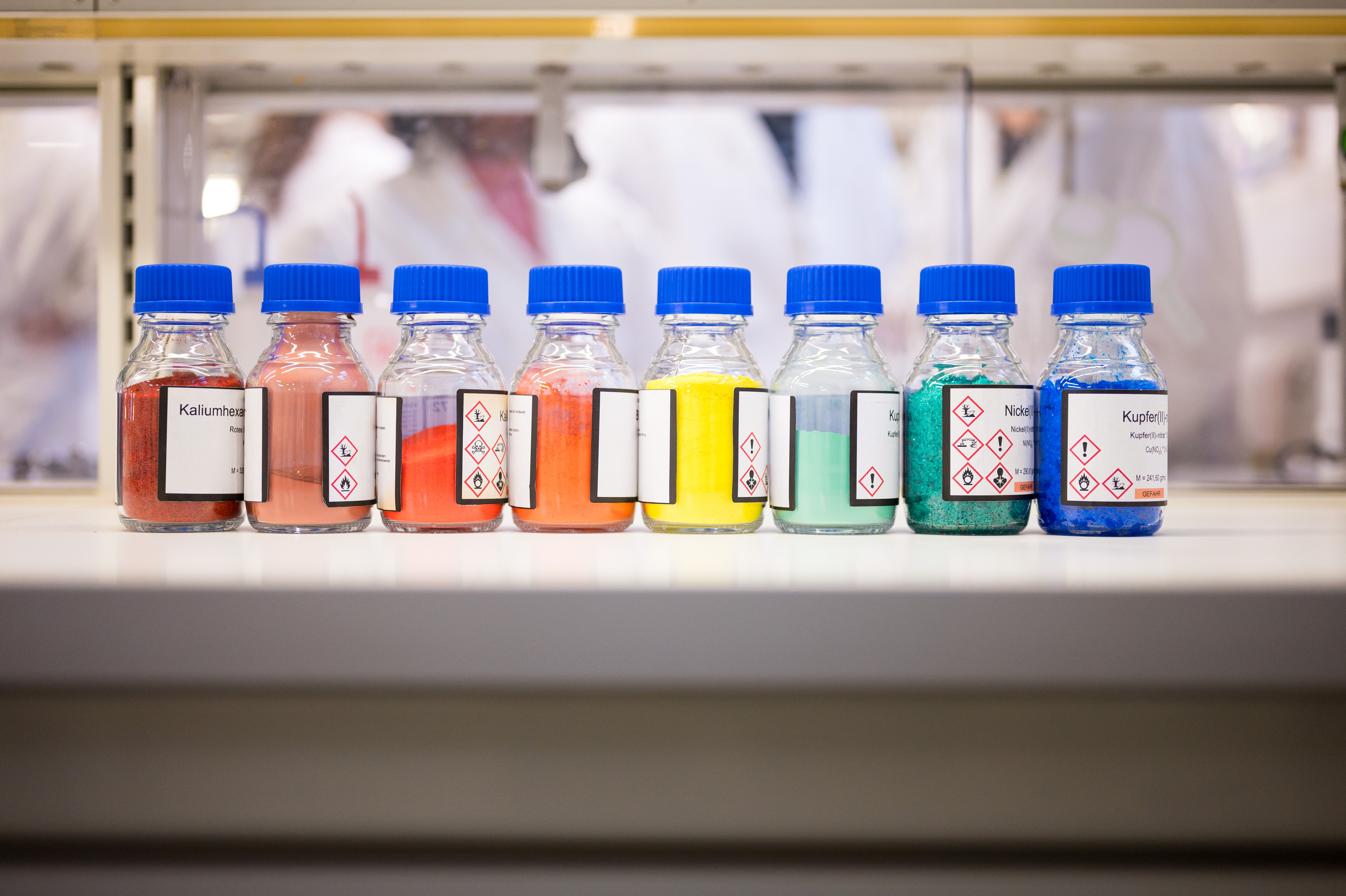 auf einer Bench (Arbeitsplatz im Labor) stehen 8 Flaschen mit blauem Deckel. In den Flaschen sind bunte Feststoff: rot, dunkelrot, 2 mal organge, gelb, türkis, grün und blau