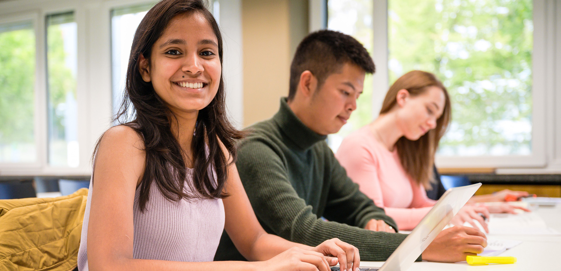 Studenten im Seminar, im Vordergrund junge Frau mit Laptop schaut in die Kamera.
